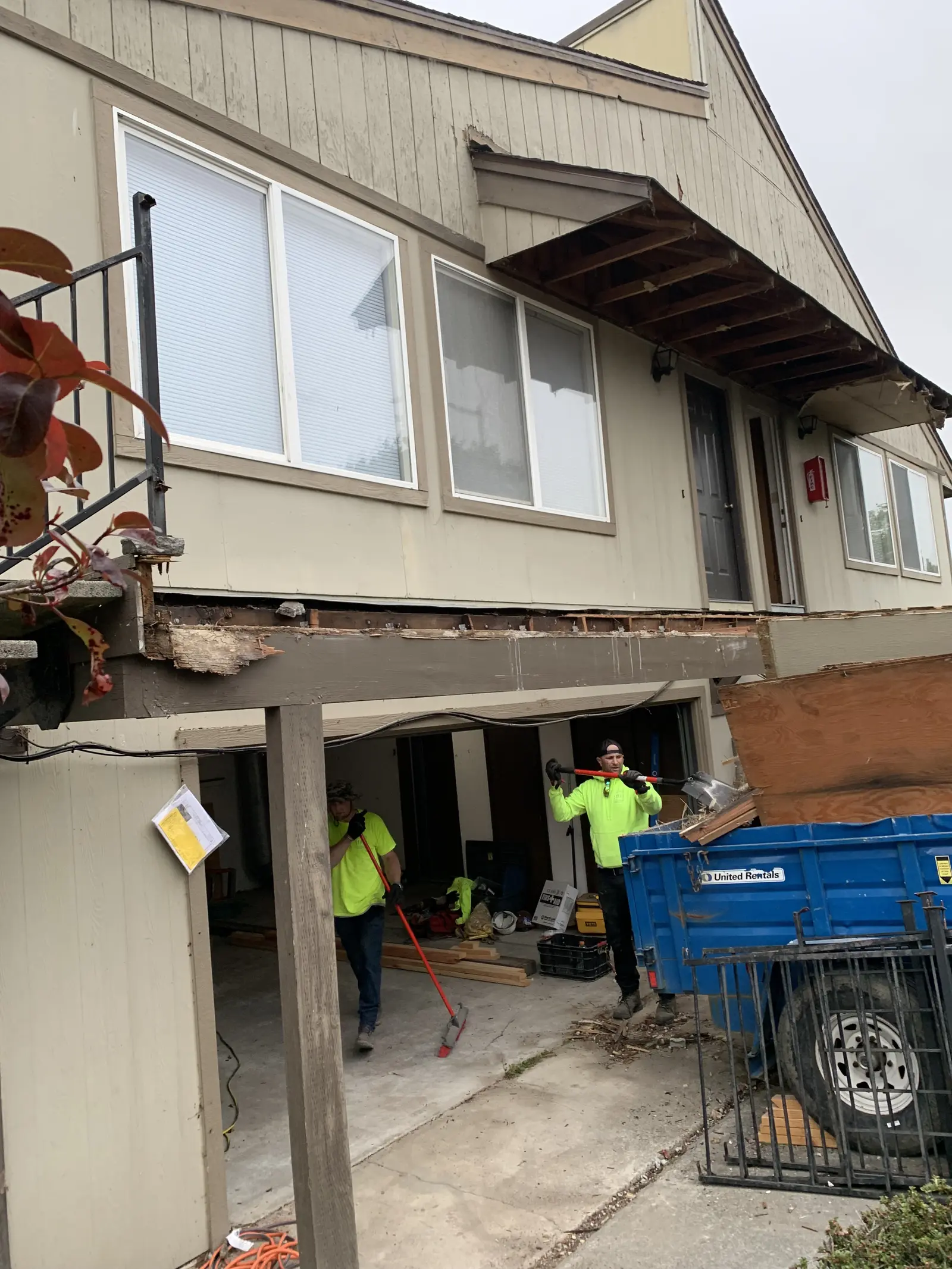 Demolition at the carport bay: rotted beam and fascia exposed over the opening, crew working, dumpster staged