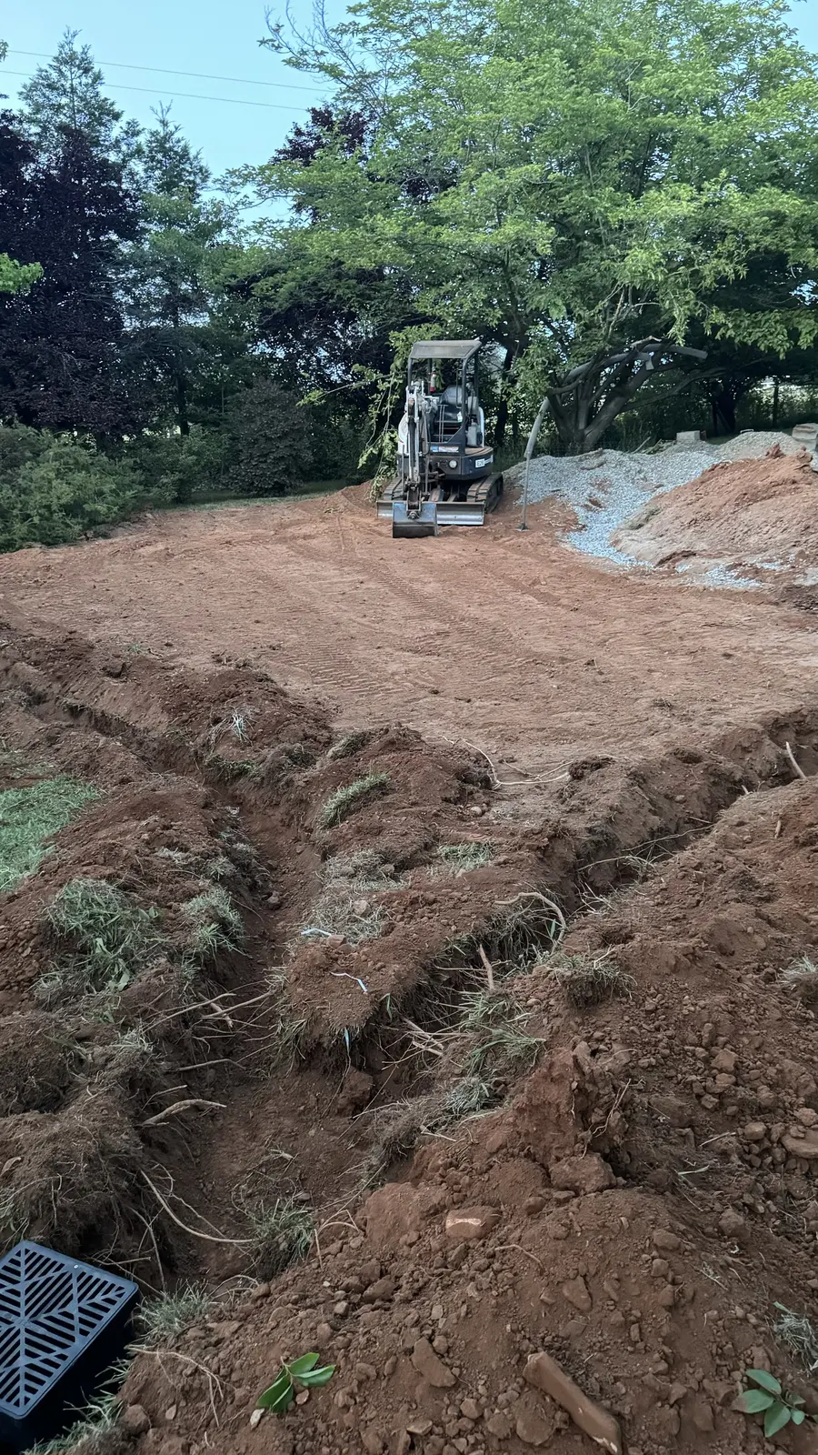 Mini excavator working the new grade with an open trench and a drainage basin grate set in the foreground