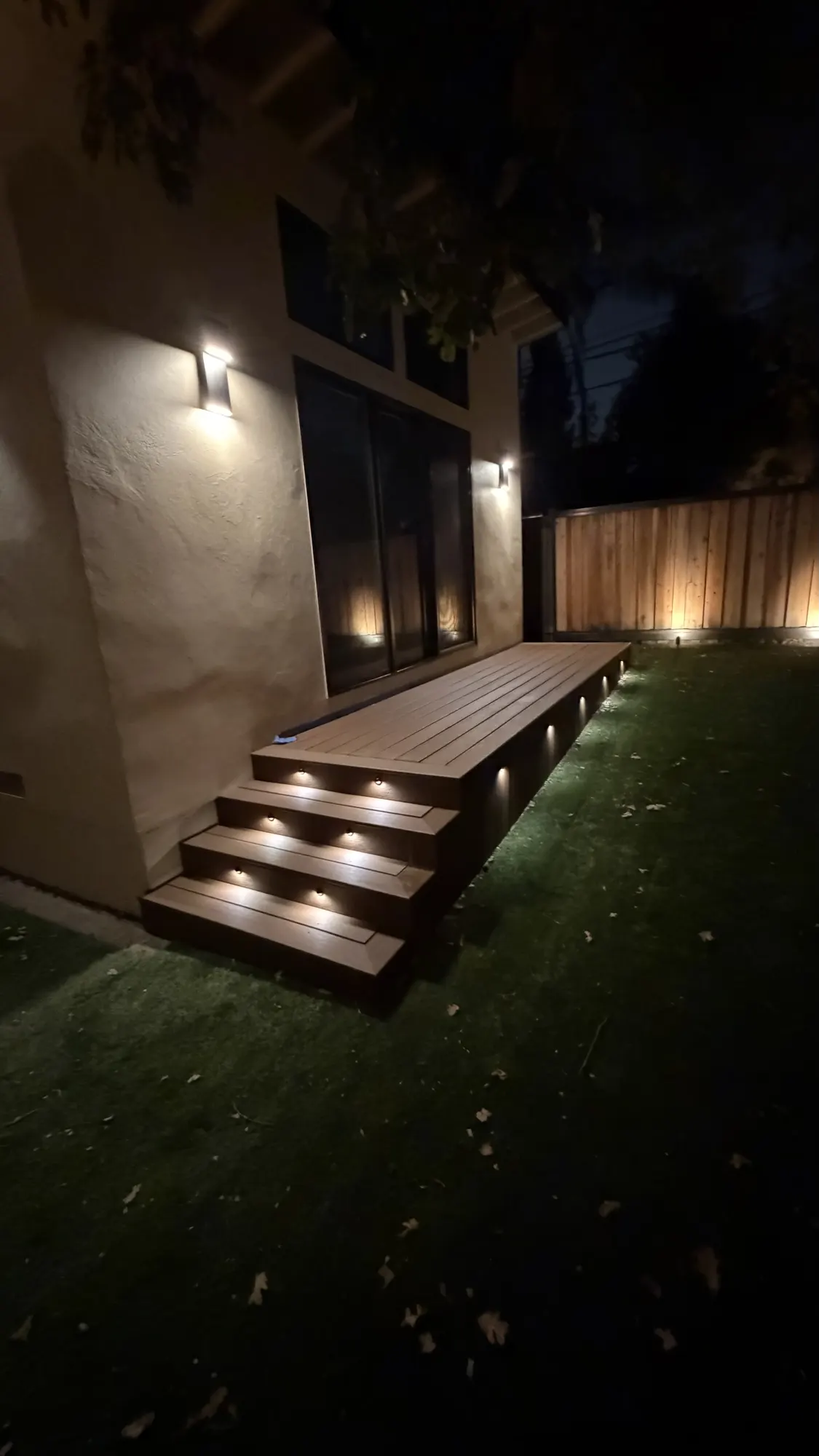 Composite deck along a stucco home at night, lit by low-voltage stair riser lights and wall sconces
